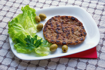 Balkan cuisine. Pljeskavica - grilled dish of minced meat - with green olives and leaves of lettuce on white plate