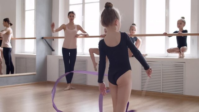 Three Caucasian girls and young teacher watching female gymnast wearing black leotard taking purple ribbon and starting exercising with it