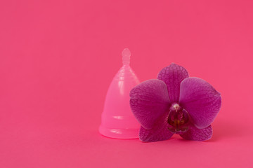 Pink menstrual cup with orchid flower on pink background. Female intimate hygiene concept. Top view