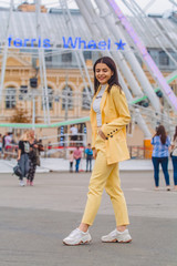 Fototapeta na wymiar Young and stylish girl walks around the city opposite the ferris wheel in yellow clasic suit. Fashion.