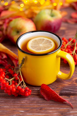 Yellow cup of tea with slice of lemon, colorful red and yellow autumn leaves, an apple  and bunch of rowanberry on wooden brown table.