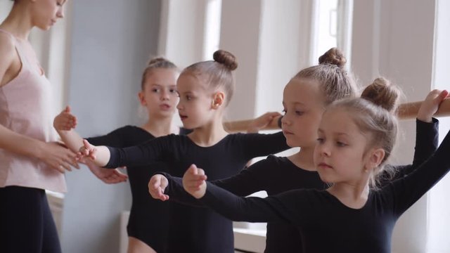 Tilt down of young Caucasian female teacher going along studio and showing little ballerinas standing in row at barre how to hold hands in right position