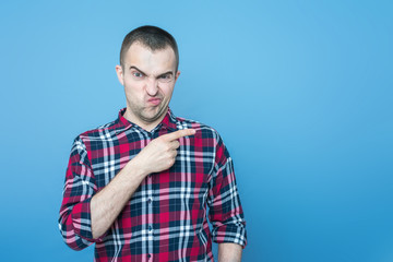 Dissatisfied young man points finger at the empty space, front view, blue, background, copy space,