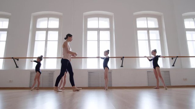 Wide shot of young professional ballerina going along studio and showing little ballerinas how to stand in right position at barre