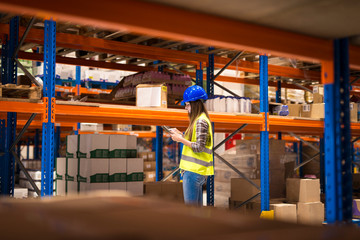Smiling warehouse worker checking packages on shelf and stock of products on her tablet computer.