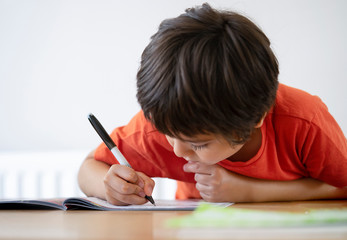 Naklejka premium School kid boy siting on table doing homework with white background, Happy Child holding black pen,Little boy is writing on white paper at the table,Elementary school and education concept