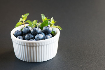 Big tasty blueberry and green mint leaves in a white plate on a dark background. Healthy berry. Vaccinium uliginosum.