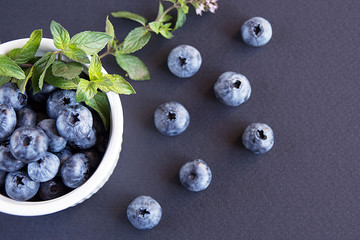 Big tasty blueberry and green mint leaves in a white plate on a dark background. Healthy berry. Vaccinium uliginosum.