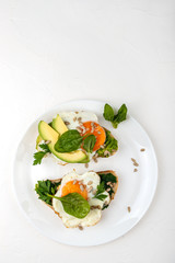 Fried eggs on a toasts bread with avocado, spinach and seeds on a white plate on the white background.