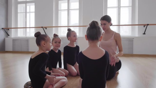 Panning of young ballet teacher sitting on floor in ballet school surrounded by Caucasian girls in black leotards and talking to them