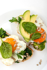 Fried eggs on a toasts bread with avocado, spinach and seeds on a white plate on the white background.