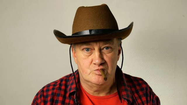 Senior man of caucasian ethnicity in a cowboy hat with a cheeky look, smokes a cigar and blows smoke towards the camera. Close-up.