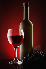 Red wine in a glass and a bottle next to grapes. Red background. Studio shoot. Close up and vertical view.
