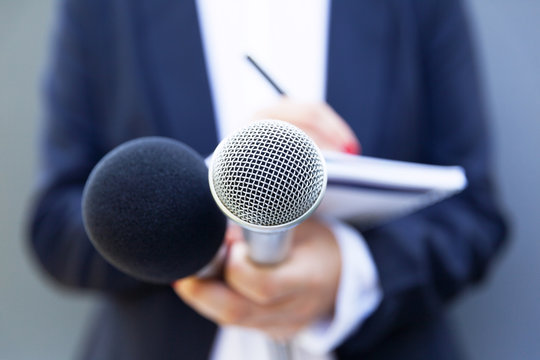 Female journalist at news conference or media event, writing notes, holding microphone