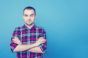 Young man in everyday clothes, he smirking, looking at camera, standing and having his arms crossed, portrait, front view, blue background, copy space