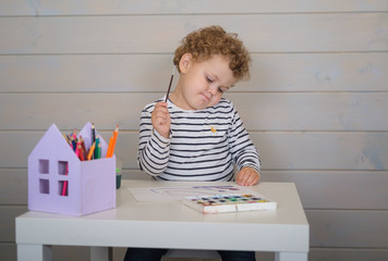 little curly boy draws watercolor paints on paper sitting at the table
