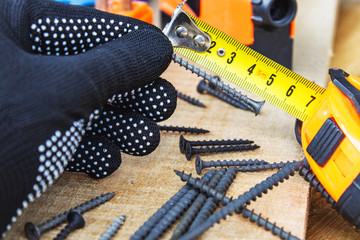 The black screw in hand and is measured by a yellow tape measure with a ruler on a wooden board...