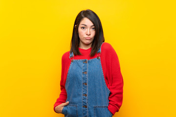 Young Mexican woman with overalls over yellow wall with sad and depressed expression