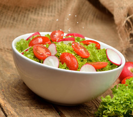 Close-up of fresh green salad Lollo Biondo with wet tomatoes and radishes with drops in a white bowl on wooden table