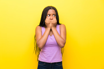 African American teenager girl with long braided hair over isolated yellow wall nervous and scared...