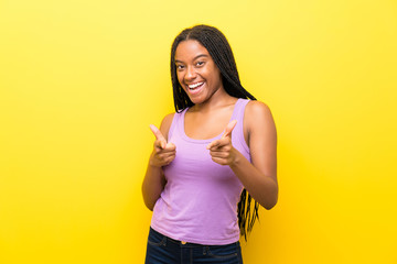 African American teenager girl with long braided hair over isolated yellow wall pointing to the front and smiling