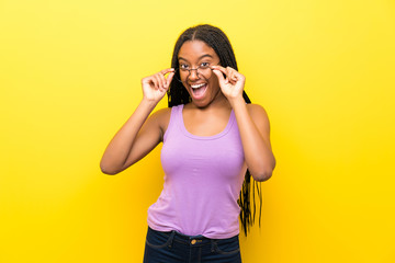 African American teenager girl with long braided hair over isolated yellow wall with glasses and surprised