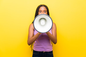 African American teenager girl with long braided hair over isolated yellow wall shouting through a megaphone