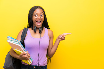 African American teenager student girl with long braided hair over isolated yellow wall surprised and pointing finger to the side - obrazy, fototapety, plakaty