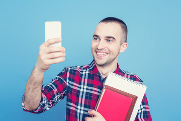 Happy student with books, smiling for selfie, using smartphone, portrait, blue background, copy...