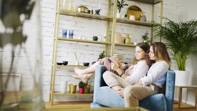 Wide view girls relaxing on blue chair in living room, discussing, reading book at background cupboard decorated colorful plates and cup. Concept LGBT couple at home