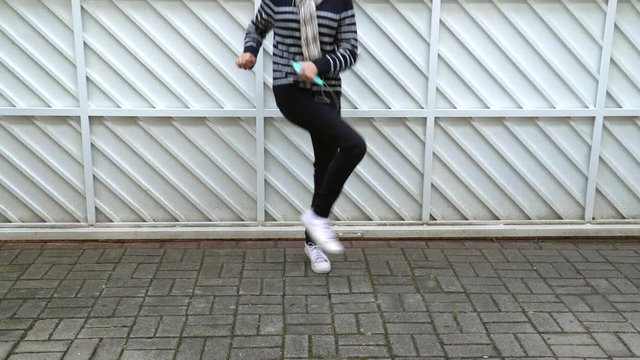 Young girl dancing and listening music on cellphone on white gate background. Unrecognizable model body holding smartphone and earphones wearing casual black pants and pullover and white sneakers.