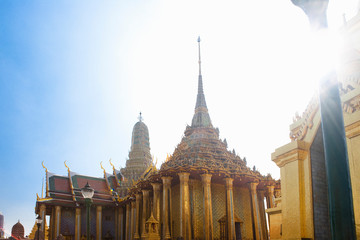 Fototapeta premium Bangkok, Thailand - A golden Buddhist temple is surrounded by columns supporting a decorative roof. A sunburst can be seen in the upper right of the shot.
