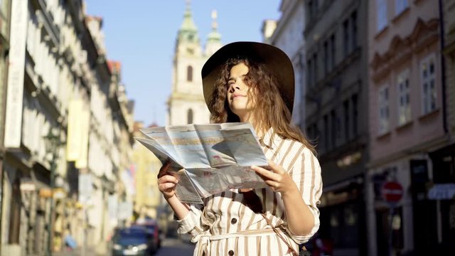 Standing young woman tourist on the street of old town Prague at morning time. Medium shot attractive girl enjoying sunny day in city, looking at map. Girl visiting the sights Praha in holiday