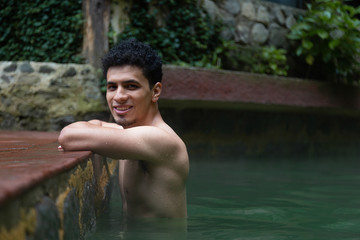 Portrait of young Hispanic man enjoying and relaxing in natural hot springs smiling and looking at camera - man at the shore of the natural spa pool