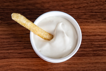 One piece of french fries in mayonnaise. Mayonnaise in small round white ceramic dish on wooden table.