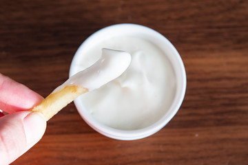 Hand holding one piece of french fries with in mayonnaise and sauce in the background. Mayonnaise in small round white ceramic dish on wooden table.