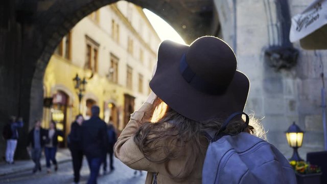 Attractive woman in hat standing on the street of old town Prague at sunset. Close-up portrait smiling girl enjoying sunny day, glance around and taking photo city sights in backlit.
