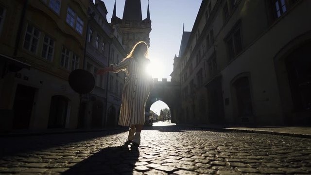 Young female tourist walking on the street of old town Prague at morning time. Girl goes toward the gamera on cobblestone pavement in backlit, enjoying sunny day and moving hands holding hat.