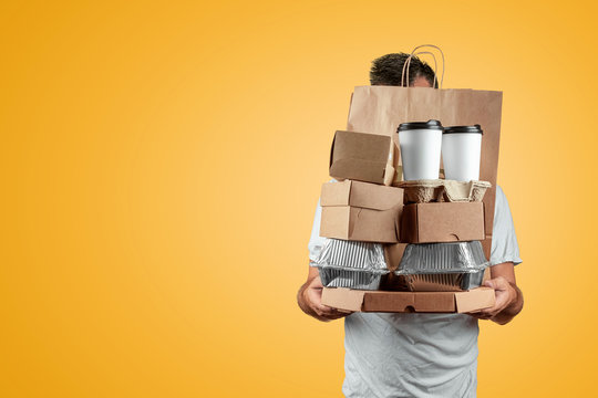 Man In A Bright T-shirt Giving A Fast Food Order Isolated On A Yellow Background. Male Worker Courier Hold A Packet Of Food. Home Delivery Of Goods From A Store Or Restaurant. Copy Space