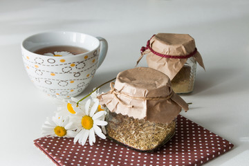 two cans closed by paper tied with a lace with tea and a cup of tea with camomile on a brown polka dot fabric on a white background