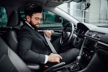 The young handsome bearded man fastening seat belt in a black business suit. A driver in a new car.