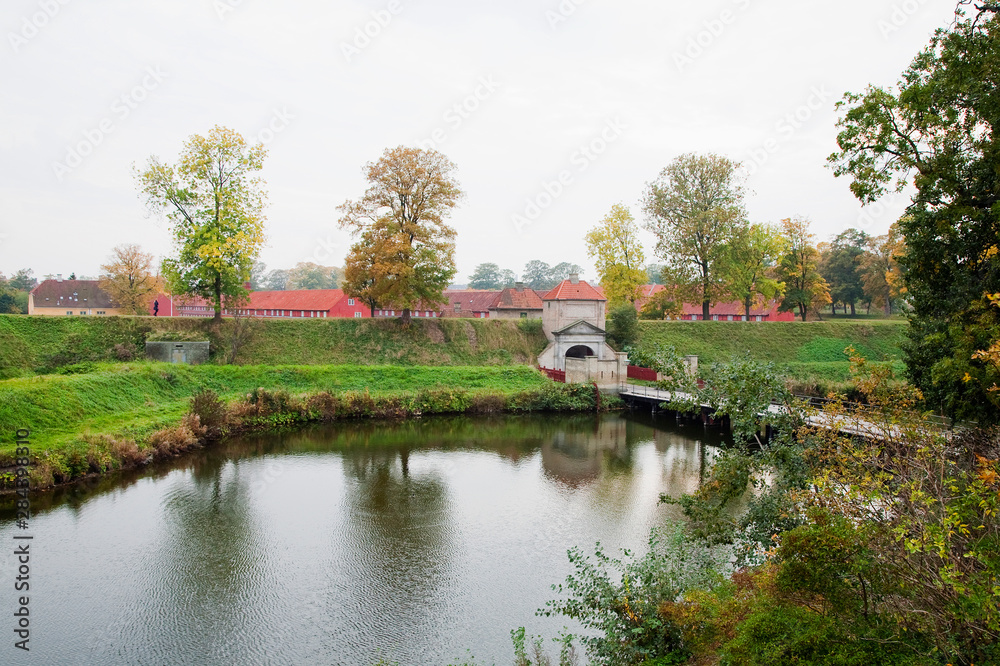 Wall mural Copenhagen, Denmark - A lake is set amidst a grassy landscape with a bridge running across it. Red army barracks can be seen in the background.