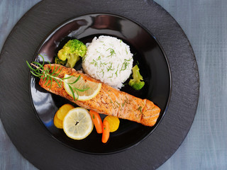 Grilled salmon fillet with rice and vegetables on black plate over black slate tray on wooden table, top view, overhead.