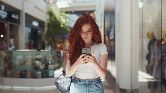 Extremely beautiful cute young redheaded student girl walking down the shopping center while actively texting via her smartphone and smiling. Modern communication, having fun, shopping time. Female