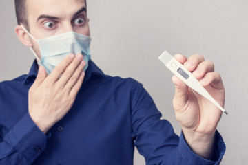Dumbfounded male in a medical mask show a thermometer in his hand, increased body temperature, close up, toned