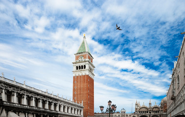 Fototapeta na wymiar Venice, Veneto, Italy - A bird is flying in the sky above a plaza in a church courtyard. A clock tower can be seen in the background.