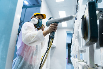 Powder coating of metal parts. A woman in a protective suit sprays white powder paint from a gun on metal products