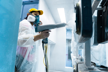 Powder coating of metal parts. A woman in a protective suit sprays white powder paint from a gun on...