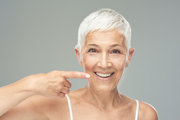 Beautiful Caucasian  smiling senior woman with short grey hair pointing at her teeth and looking at...
