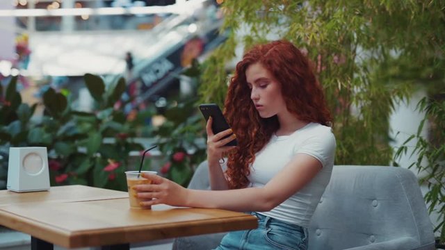 Attractive young ginger hipster girl in casual outfit taking photo of coffee via modern smartphone to share photos with followers in social networks. While sitting in coffee and waiting for his friend